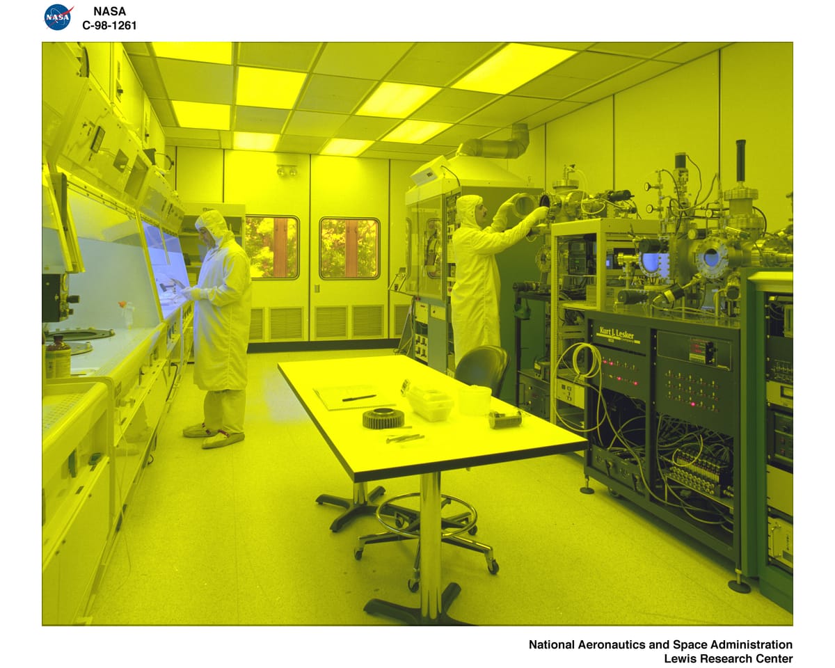 NASA Glenn cleanroom technicians in full protective garments working beneath amber lighting around semiconductor process equipment, illustrating the controlled manufacturing environment behind advanced chips.